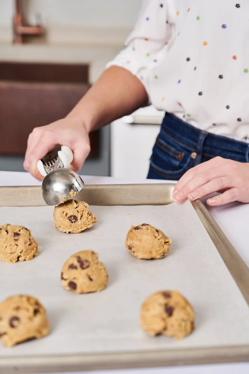 Hands using a stainless steel cookie scoop to place dough on a parchment-lined baking sheet
