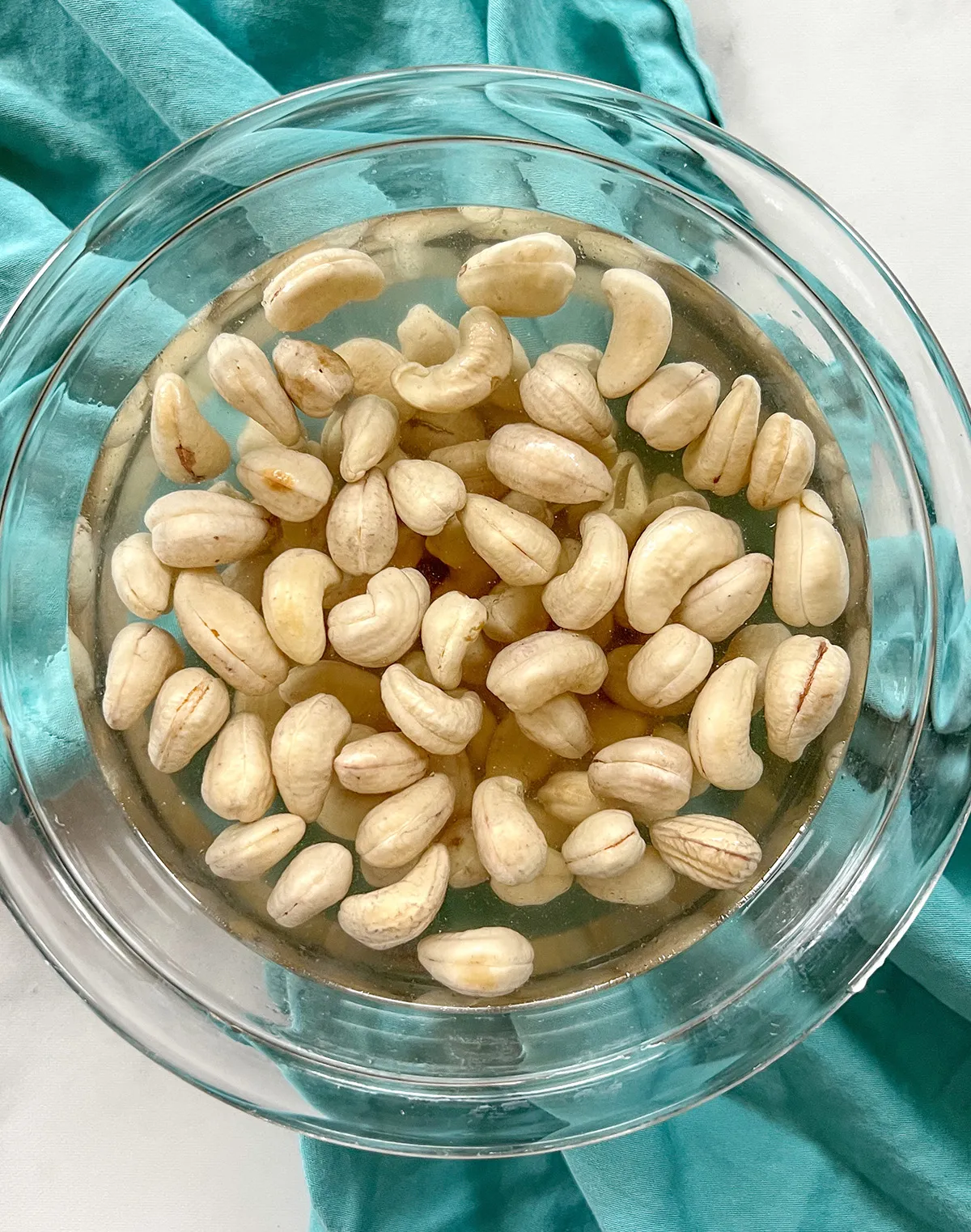 Close up of raw cashews soaking in a bowl of water next to fresh garlic and limes